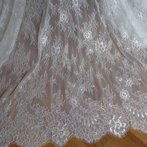 May include: White lace fabric with a delicate floral pattern. The lace has a scalloped edge.