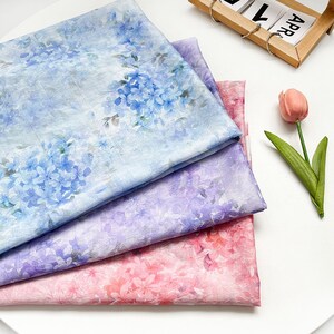 May include: Three folded pieces of fabric featuring a floral hydrangea pattern. The top layer is light blue, the middle layer is purple, and the bottom layer is pink. Each piece displays a repeating print of hydrangea blossoms and green leaves.