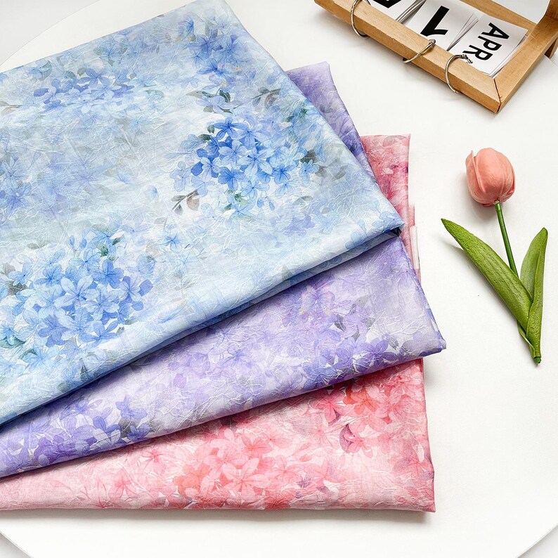 May include: Three folded pieces of fabric featuring a floral hydrangea pattern. The top layer is light blue, the middle layer is purple, and the bottom layer is pink. Each piece displays a repeating print of hydrangea blossoms and green leaves.