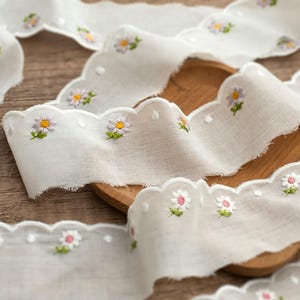 May include: A white fabric ribbon featuring a scalloped edge and embroidered daisy motifs in shades of purple, yellow, and pink with green stems. The ribbon has a semi-sheer texture and frayed edges, displaying a repeating floral pattern throughout.