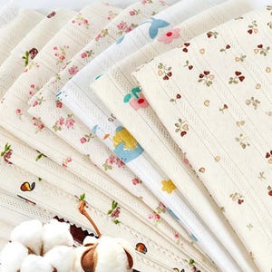 May include: A collection of folded fabric swatches featuring various patterns including small floral prints, cherries, and hearts on cream-colored backgrounds. The fabrics have a textured, ribbed appearance and are arranged in a fan-like display.