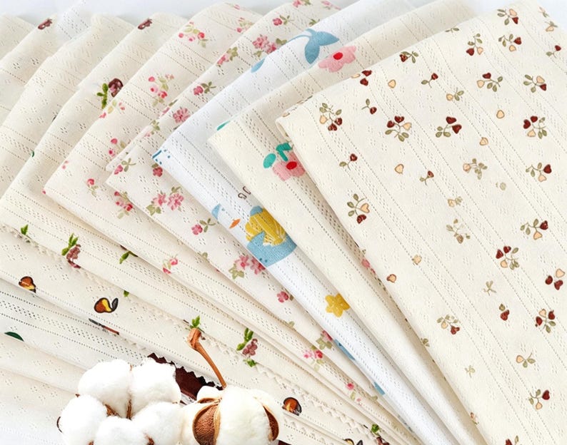 May include: A collection of folded fabric swatches featuring various patterns including small floral prints, cherries, and hearts on cream-colored backgrounds. The fabrics have a textured, ribbed appearance and are arranged in a fan-like display.
