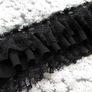 May include: Black lace trim with multiple layers of ruffles. The trim is on a white background with a floral pattern.