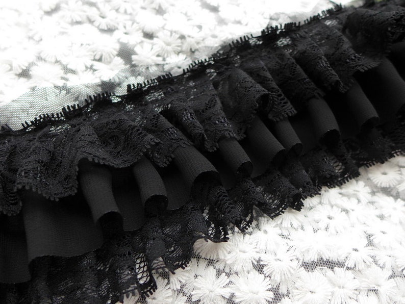 May include: Black lace trim with multiple layers of ruffles. The trim is on a white background with a floral pattern.