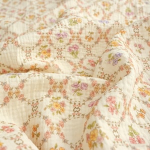 May include: Cream-colored fabric with a floral pattern. The design features a repeating pattern of flowers in shades of pink, yellow, and purple, arranged in a circular pattern. The fabric has a soft, textured appearance, suggesting a lightweight material.