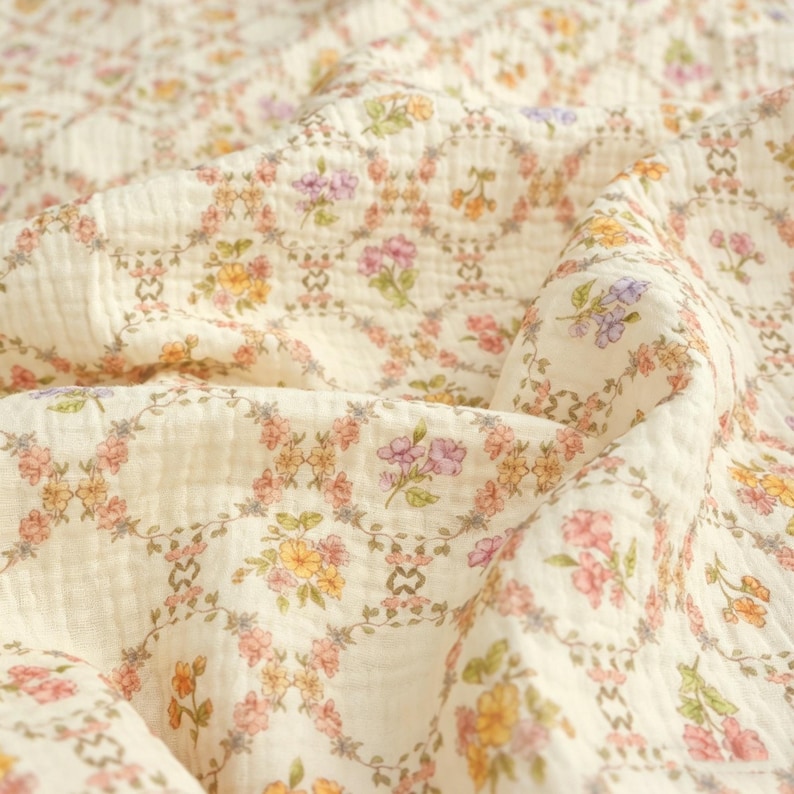 May include: Cream-colored fabric with a floral pattern. The design features a repeating pattern of flowers in shades of pink, yellow, and purple, arranged in a circular pattern. The fabric has a soft, textured appearance, suggesting a lightweight material.