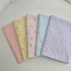 May include: Five folded fabric pieces in pastel colors: pink, yellow, pink, light blue, and lavender. Each fabric has a gingham pattern with small pink bow accents. The fabrics are arranged in a fan shape, showcasing the variety of colors and patterns.