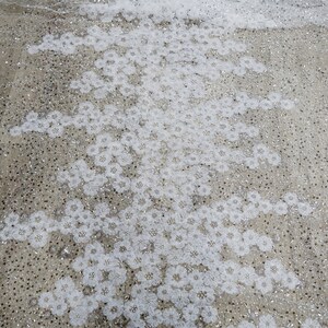 以下が含まれることがあります： 繊細な花柄と輝く装飾が施された白いチュール生地。生地は透き通っており、柔らかく流れるような質感です。