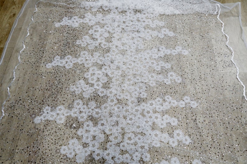 以下が含まれることがあります： 繊細な花柄と輝く装飾が施された白いチュール生地。生地は透き通っており、柔らかく流れるような質感です。