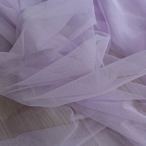 May include: A close-up of a soft, light purple tulle fabric. The fabric is delicate and sheer, with a fine mesh texture.