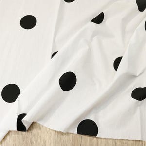 May include: A white fabric featuring a repeating pattern of large, solid black polka dots. The material is draped to show folds and texture, with visible raw edges along the bottom hem, displaying a classic high-contrast monochrome design.
