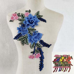 May include: A floral embroidery patch featuring large blue roses, smaller pink blossoms, and green leaves with dark blue accents. The design is arranged in an asymmetrical cluster with trailing stems and foliage, displayed on a neutral mannequin torso.