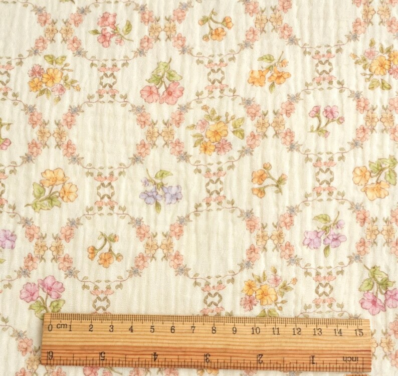 May include: Cream-colored fabric with a floral pattern. The design features oval frames with pink, yellow, and purple flowers, connected by a delicate vine-like pattern. A wooden ruler shows measurements up to 15 inches.