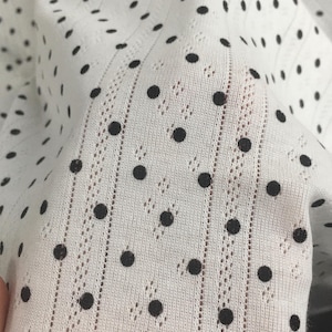 May include: A white fabric featuring a pattern of small black polka dots. The material has a vertical ribbed texture with delicate, openwork eyelet detailing running in parallel lines between the rows of dots, creating a lightweight appearance.