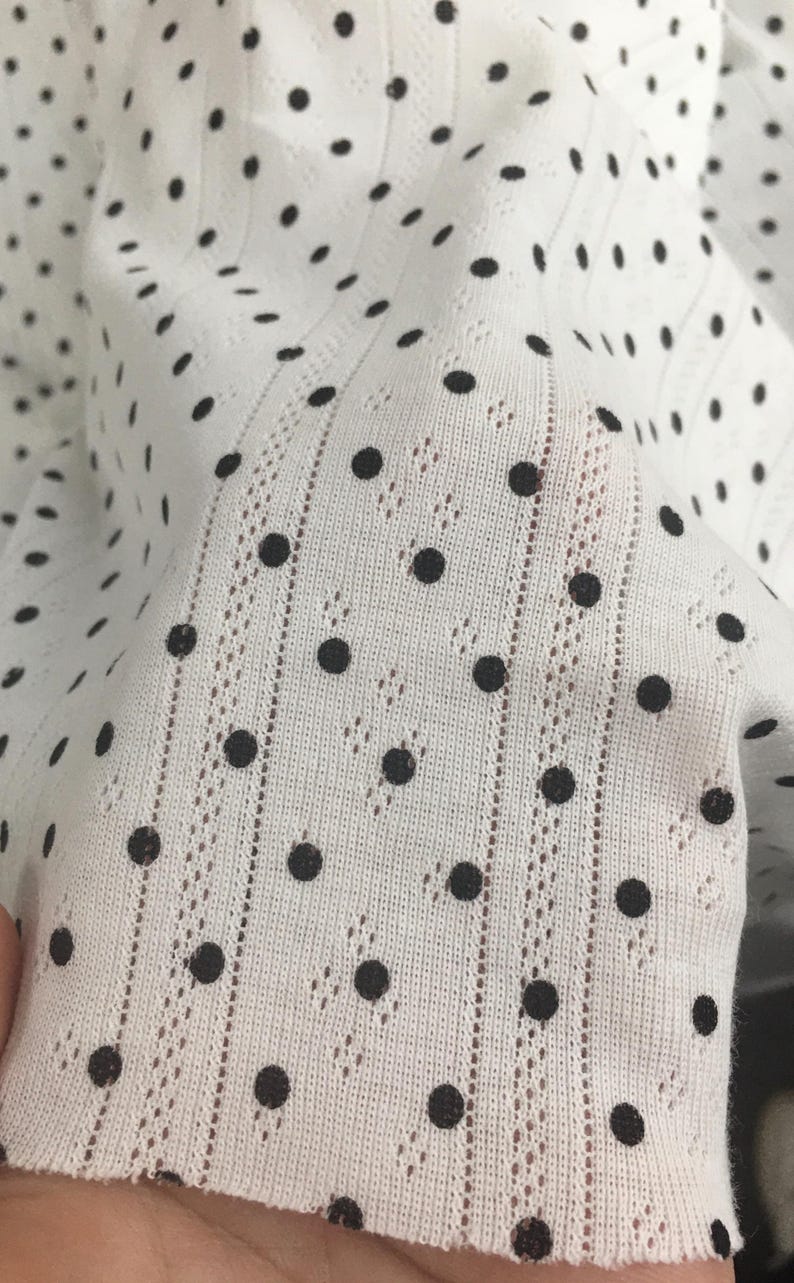 May include: A white fabric featuring a pattern of small black polka dots. The material has a vertical ribbed texture with delicate, openwork eyelet detailing running in parallel lines between the rows of dots, creating a lightweight appearance.