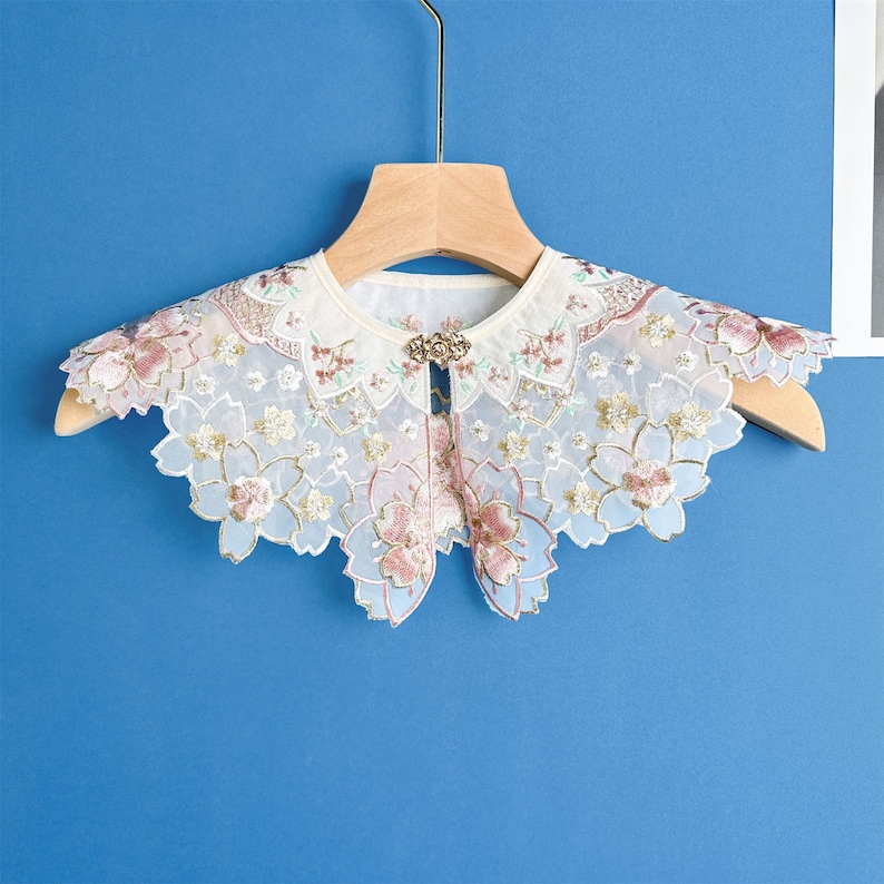 May include: A white lace collar with floral embroidery and a gold button detail. The collar is on a wooden hanger against a blue background.