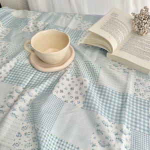 May include: A cream-coloured teacup and saucer rest on a patchwork quilt with blue and white floral and gingham patterns. An open book and small bouquet of dried flowers are also on the quilt, creating a cosy, inviting scene.
