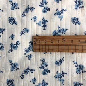 May include: Close-up of a cream-colored fabric with a ribbed texture and small blue floral patterns. A wooden ruler is placed on the fabric, showing measurements in centimeters.