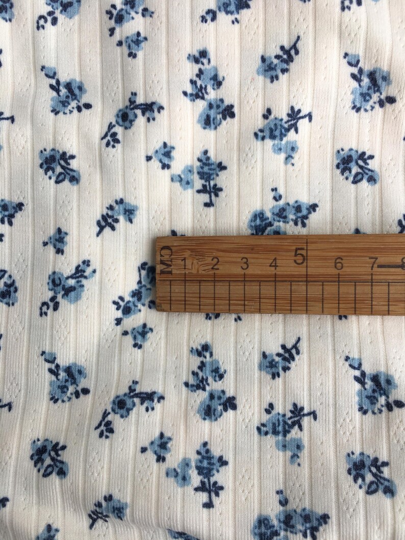 May include: Close-up of a cream-colored fabric with a ribbed texture and small blue floral patterns. A wooden ruler is placed on the fabric, showing measurements in centimeters.