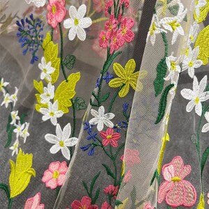 May include: A sheer fabric with embroidered floral designs in shades of pink, white, yellow, and green. The fabric has a delicate, airy feel and is perfect for creating garments or accessories.