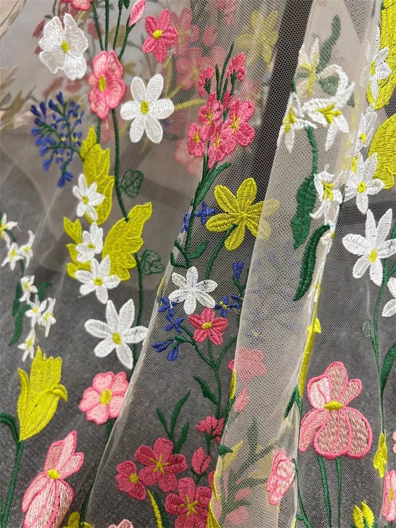 May include: A sheer fabric with embroidered floral designs in shades of pink, white, yellow, and green. The fabric has a delicate, airy feel and is perfect for creating garments or accessories.