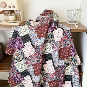 Puede incluir: Una colcha de patchwork que presenta una variedad de bloques de tela cuadrados con diversos patrones florales en tonos burdeos, azul marino, azul claro, crema y verde. La colcha est&aacute; colocada sobre una superficie de madera con un dise&ntilde;o de rejilla.