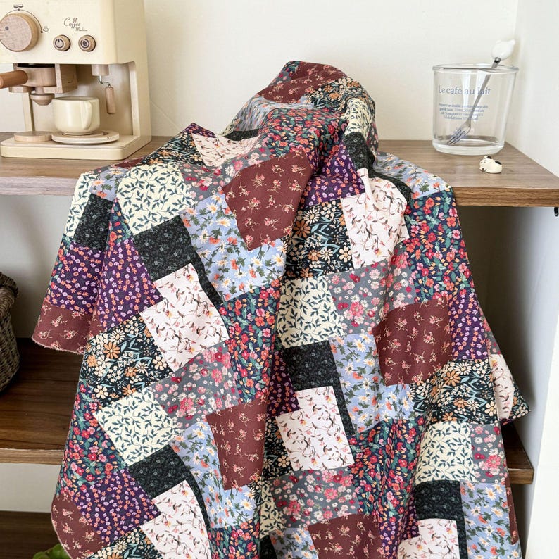 Puede incluir: Una colcha de patchwork que presenta una variedad de bloques de tela cuadrados con diversos patrones florales en tonos burdeos, azul marino, azul claro, crema y verde. La colcha est&aacute; colocada sobre una superficie de madera con un dise&ntilde;o de rejilla.
