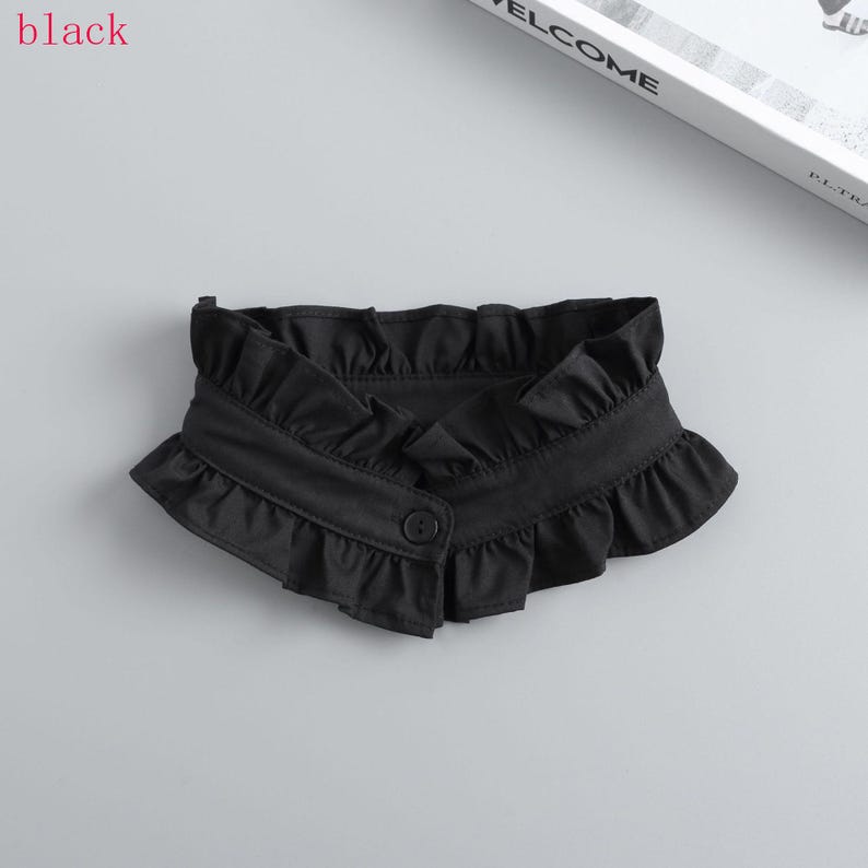 May include: A black ruffled collar with a button closure. The collar is made of a soft fabric and features a decorative ruffle along the edges. The word "black" is in the top left corner.