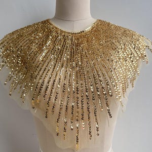 Może przedstawiać: Złoty kołnierz z cekinami, promieniujący od dekoltu. Kołnierz wykonany jest z przezroczystej, beżowej tkaniny i gęsto pokryty małymi, okrągłymi, złotymi cekinami. Idealny do dekoracji odzieży.