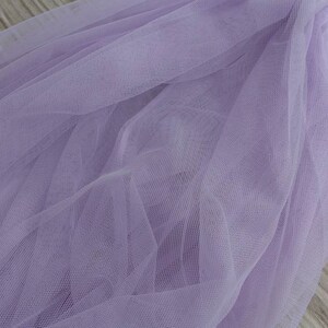 May include: A piece of soft, lilac tulle fabric with a delicate, fine mesh texture. The fabric is draped and folded, creating a soft, flowing look.
