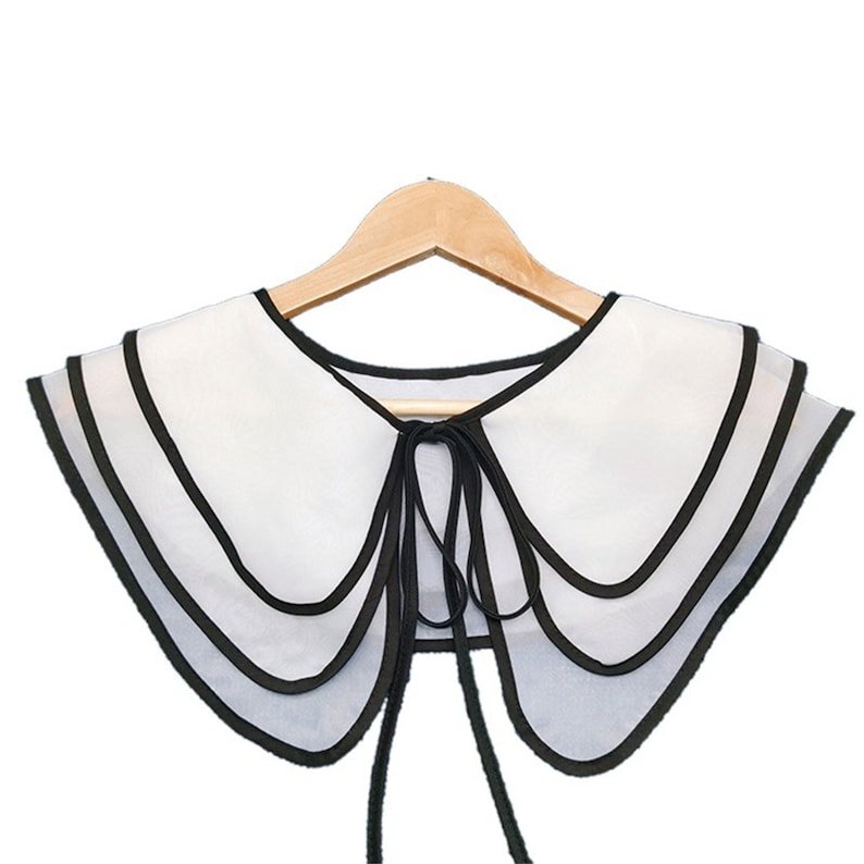 May include: A white layered collar with black trim and a black ribbon tie. The collar is on a wooden hanger.