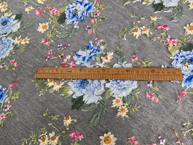 Puede incluir: Tela gris con un patr&oacute;n floral con flores azules, blancas y rosas. Una regla de madera con marcas en pulgadas se coloca sobre la tela.