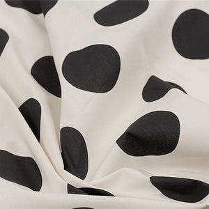 May include: A fabric featuring a pattern of irregular black spots scattered across an off-white background. The material is shown with soft folds and drapes, highlighting the contrast between the dark organic shapes and the light base color.