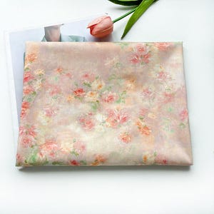 May include: A folded fabric with a floral pattern featuring pink, peach, and white roses. The fabric has a soft, muted color palette and a watercolor-like effect. A pink tulip and green stem are in the upper right corner.