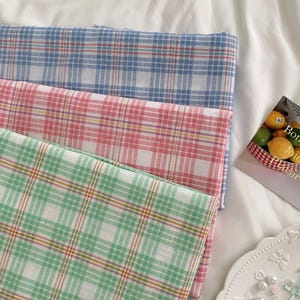 May include: Three folded fabric squares in blue, pink, and green plaid patterns. The fabrics are stacked on a white surface. The plaid design features thin lines in contrasting colors, creating a classic, timeless look. The fabrics appear to be lightweight.
