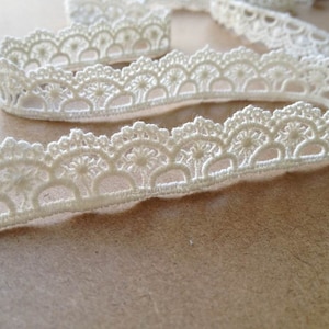 May include: Close-up of ivory-colored lace trim with a scalloped edge. The lace features a repeating floral pattern and is arranged in a flowing, ribbon-like form. The trim is likely used for sewing, crafting, or adding decorative detail to garments or home decor.
