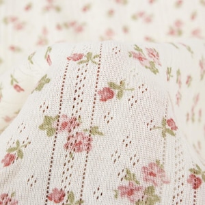 May include: A cream-colored knit fabric featuring a repeating pattern of small pink rosebuds with green leaves. The textile includes vertical rows of delicate eyelet lace detailing, creating a textured, lightweight appearance throughout the material.