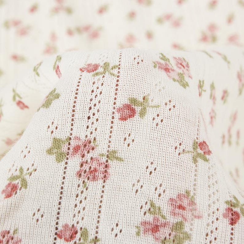 May include: A cream-colored knit fabric featuring a repeating pattern of small pink rosebuds with green leaves. The textile includes vertical rows of delicate eyelet lace detailing, creating a textured, lightweight appearance throughout the material.