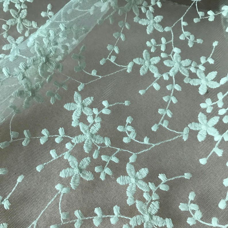 K&ouml;nnte beinhalten: Blassgr&uuml;ne florale Stickerei auf einem durchsichtigen wei&szlig;en Stoff. Das Design zeigt zarte Zweige mit kleinen Bl&uuml;ten.
