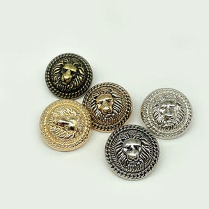 May include: Five metal buttons with lion head designs. The buttons are in various metallic finishes, including gold, bronze, and silver. Each button features a detailed lion's head surrounded by a rope-like border. These buttons are suitable for coats, jackets, or other garments.