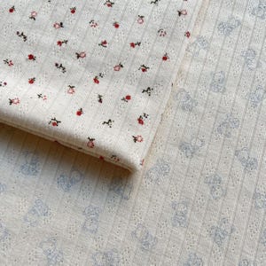 May include: Close-up of two pieces of cream-colored fabric. One piece is folded, showing a pattern of small red and pink flowers. The other piece has a pattern of light blue teddy bears.