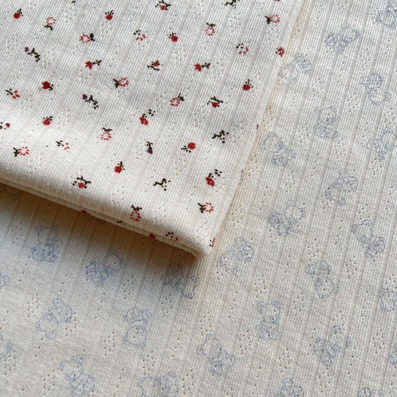 May include: Close-up of two pieces of cream-colored fabric. One piece is folded, showing a pattern of small red and pink flowers. The other piece has a pattern of light blue teddy bears.