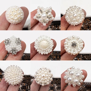 May include: A collection of nine decorative buttons featuring various arrangements of white faux pearls, clear rhinestones, and sequins. Each button displays a unique circular or star-shaped pattern, combining glossy pearl beads with sparkling accents.