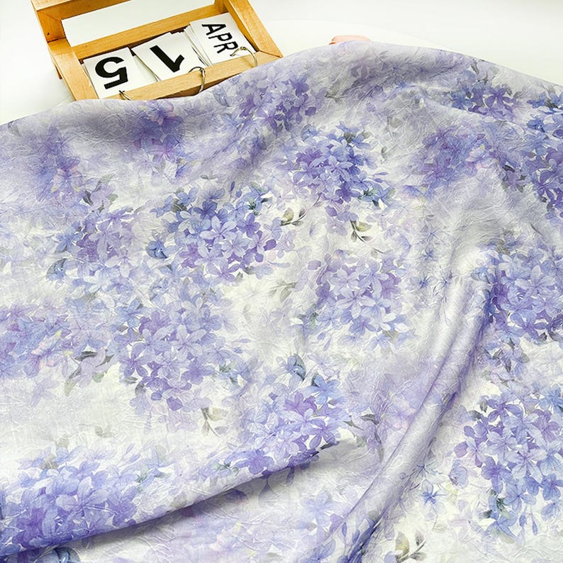 May include: A lightweight fabric featuring a repeating pattern of purple and light blue hydrangea flower clusters on a white background. The material has a slightly crinkled texture and is draped to show folds across the surface of the textile.