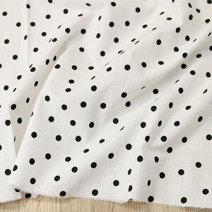 May include: A white fabric featuring a repeating pattern of solid black polka dots. The material is draped in soft folds, highlighting the texture of the textile and the consistent size and spacing of the circular black spots across the surface.