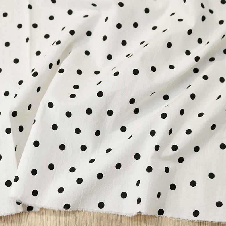May include: A white fabric featuring a repeating pattern of solid black polka dots. The material is draped in soft folds, highlighting the texture of the textile and the consistent size and spacing of the circular black spots across the surface.