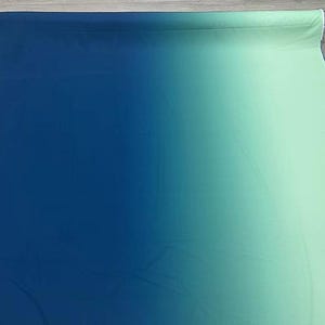 Puede incluir: Un rollo de tela con un diseño degradado, que transiciona de azul oscuro a un verde agua claro. La tela es suave y parece ser un material ligero, adecuado para proyectos de ropa o decoración del hogar.