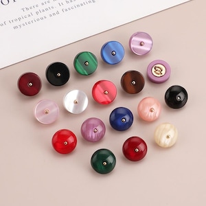 Puede incluir: Una colección de coloridos pomos redondos para armarios en varios tonos, incluyendo rojo, verde, azul y rosa. Cada pomo tiene un pequeño centro dorado. Los pomos están dispuestos sobre una superficie rosa claro.