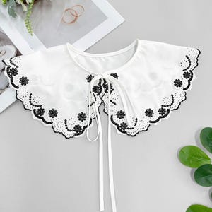 May include: A white, embroidered collar with a scalloped edge and black floral detailing. The collar has a tie closure at the front and is likely an accessory for blouses or dresses.