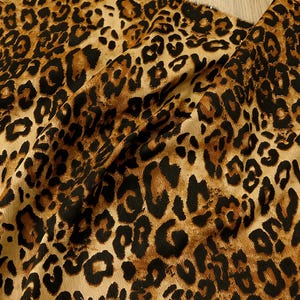 Puede incluir: Primer plano de una tela con estampado de leopardo. La tela presenta un fondo beige con manchas marrones oscuras y negras. La tela está doblada, mostrando la textura y la caída del material.